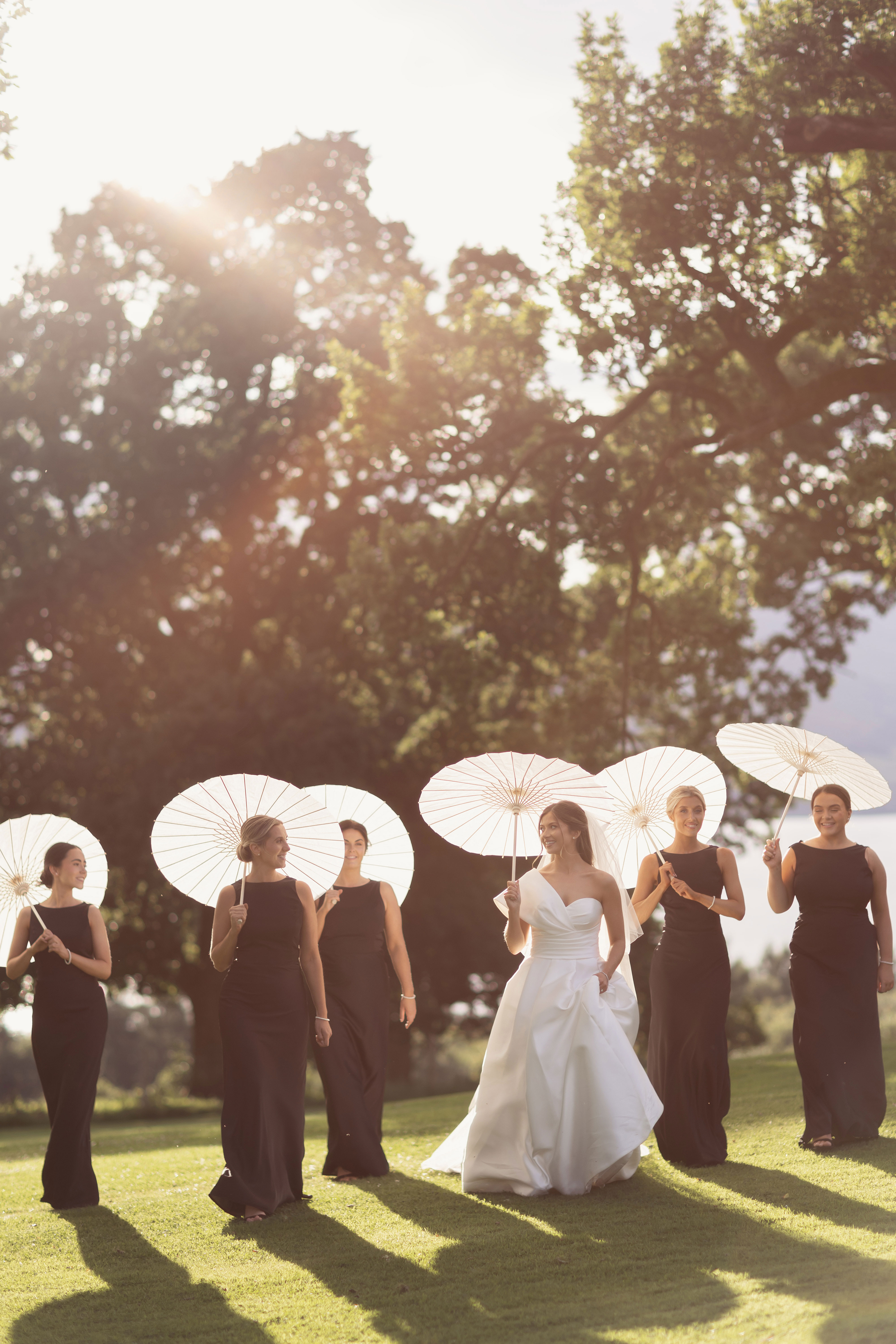 Bride and bridesmaids walking across a sunlit lawn with white parasols during golden hour at a Scottish wedding venue.