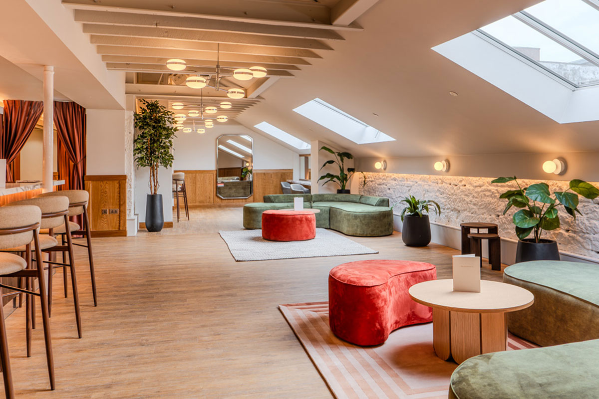 cosy lit room has wood floors, sloped ceilings, cushioned seating in green and red-orange colours with plants, mirrors and bar stool seating scattered throughout