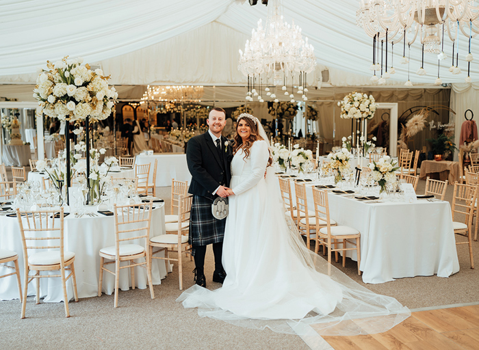 A man in a dark kilt holding hands with a bride in a long sleeved wedding dress as they stand inside a marquee with lots of tables, chairs, white flowers and chandeliers 