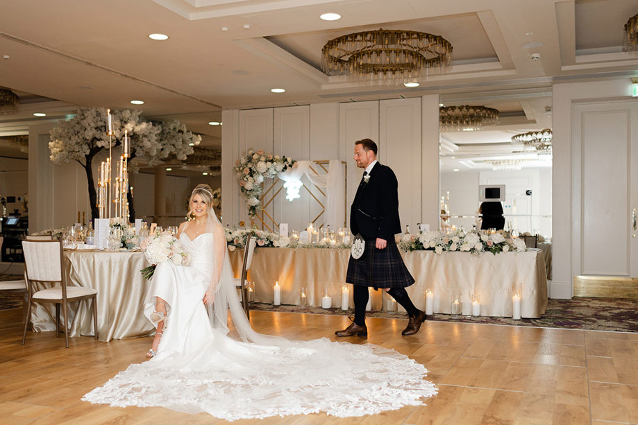 a seated bride in a room set for a wedding dinner, with a groom in a kilt walking towards her.