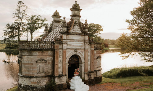 A bride wearing a wedding dress with a voluminous tulle skirt standing next to a groom wearing green stand in the door way of a small building in front of a large pond