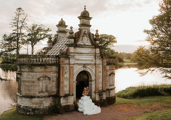 A bride wearing a wedding dress with a voluminous tulle skirt standing next to a groom wearing green stand in the door way of a small building in front of a large pond