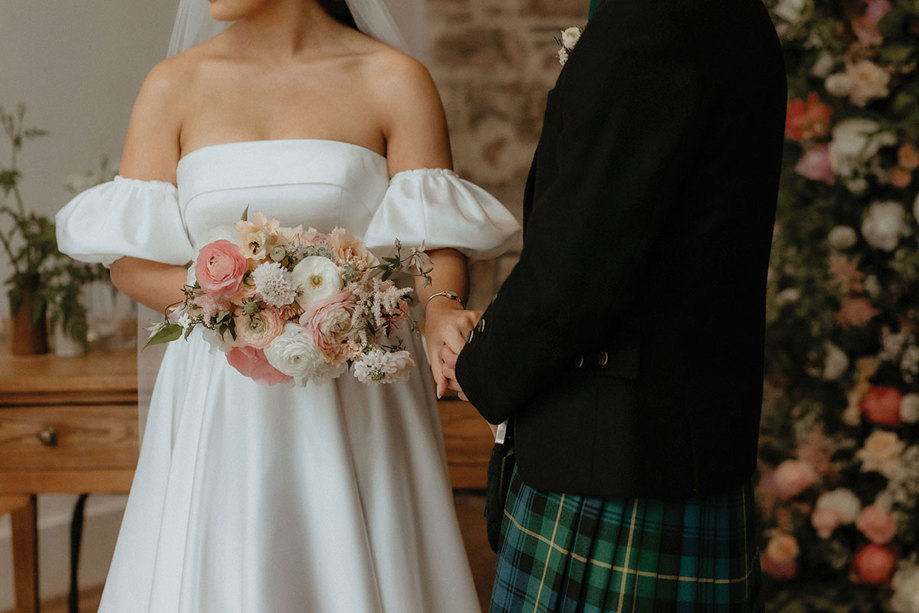 bride in off-the-shoulder wedding dress holds a polished bouquet while also holding hands with the groom