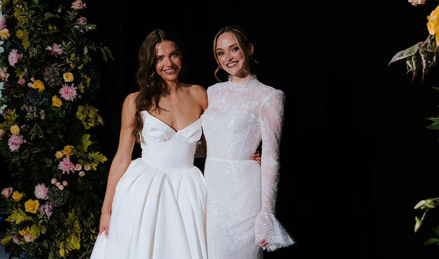 Two models wearing modern white bridal gowns posed between floral installations at The Scottish Wedding Show.