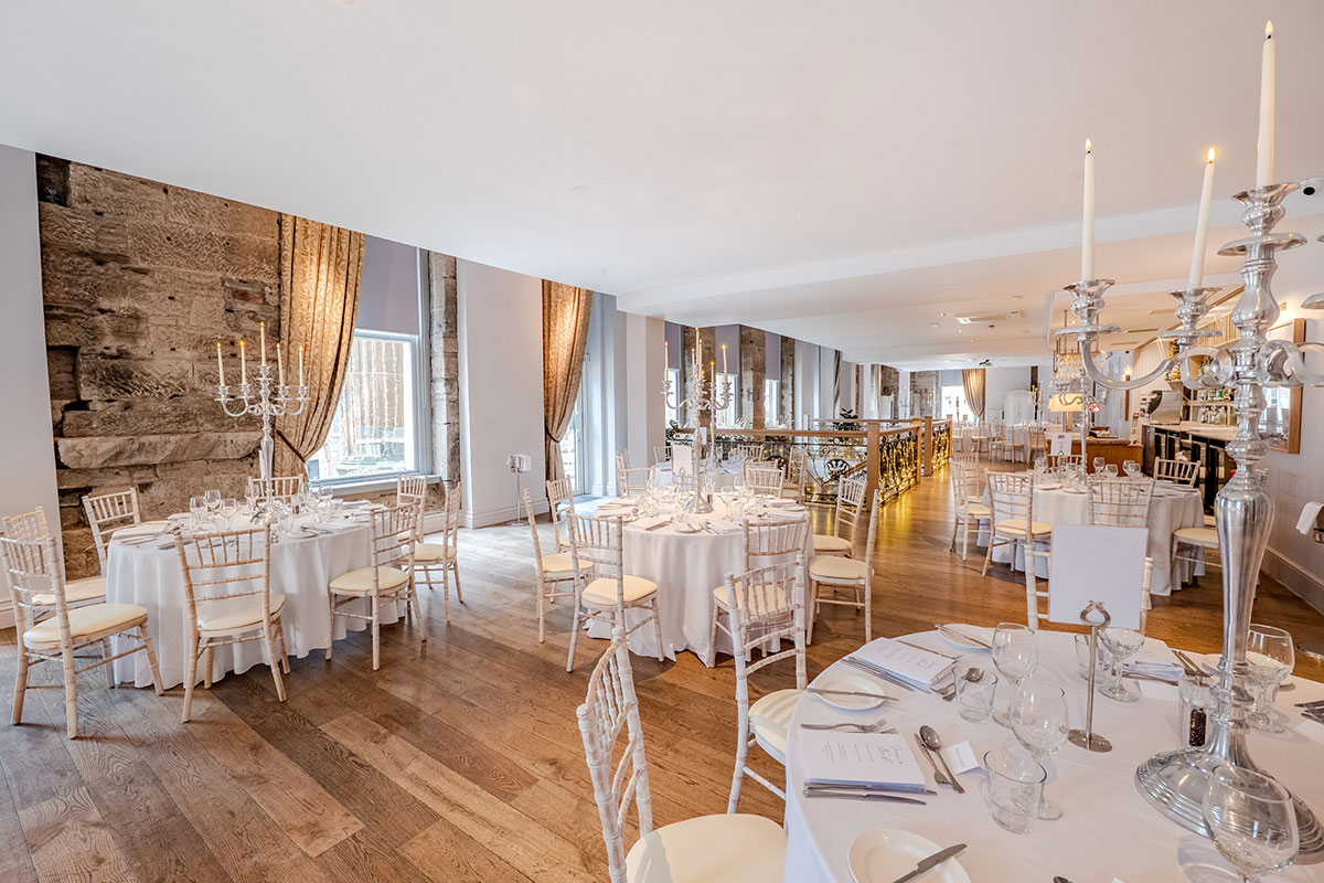 a large room with wooden floors has several round tables set with white table cloths glassware, silverware and candelabras