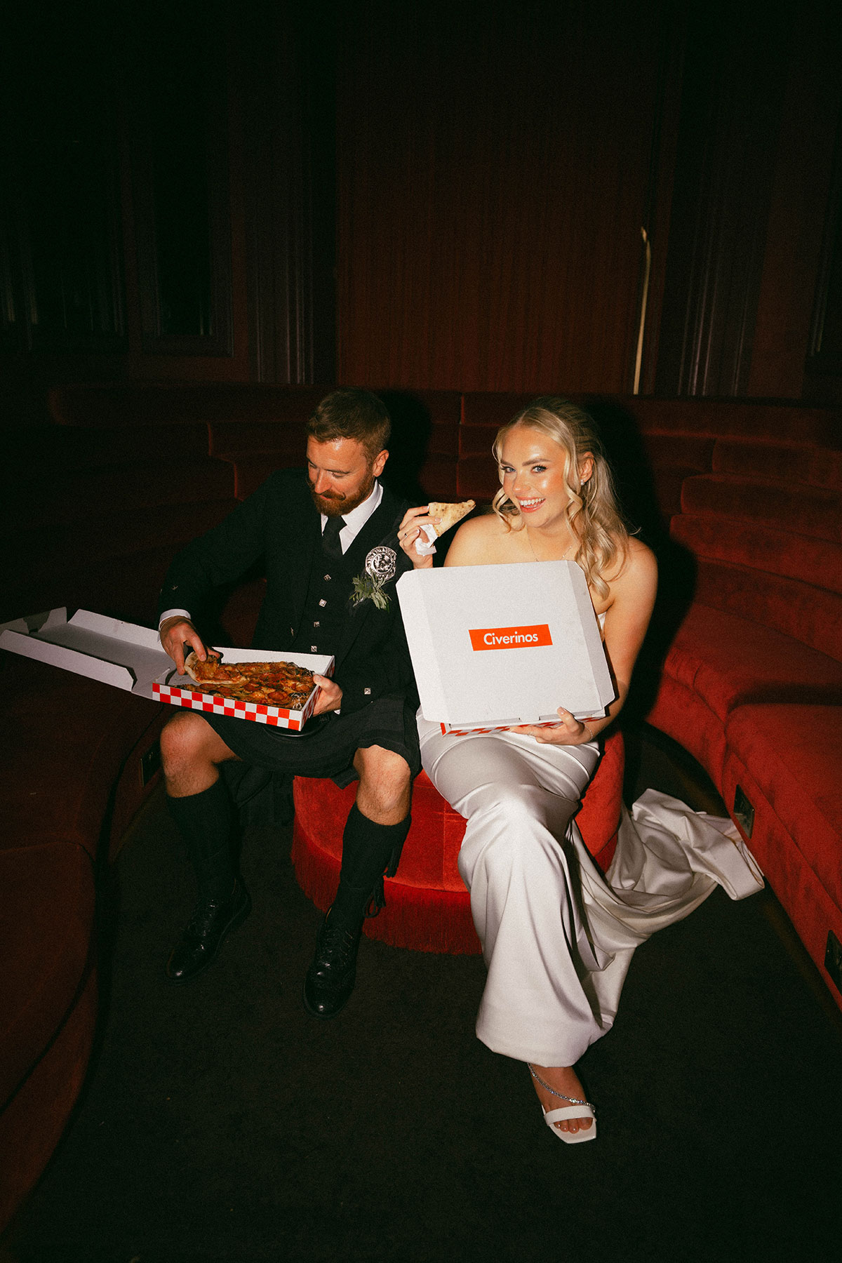 bride and groom enjoying pizza together on red velvet seating at wedding reception