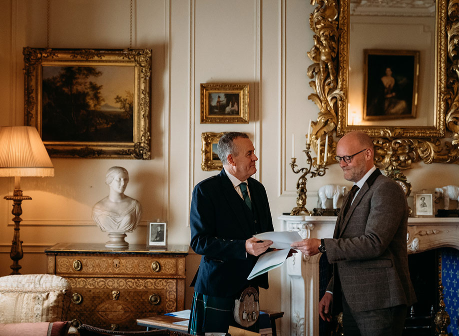 two men standing in a grand room filled with antique furniture and gold framed paintings, one passes paperwork to the other