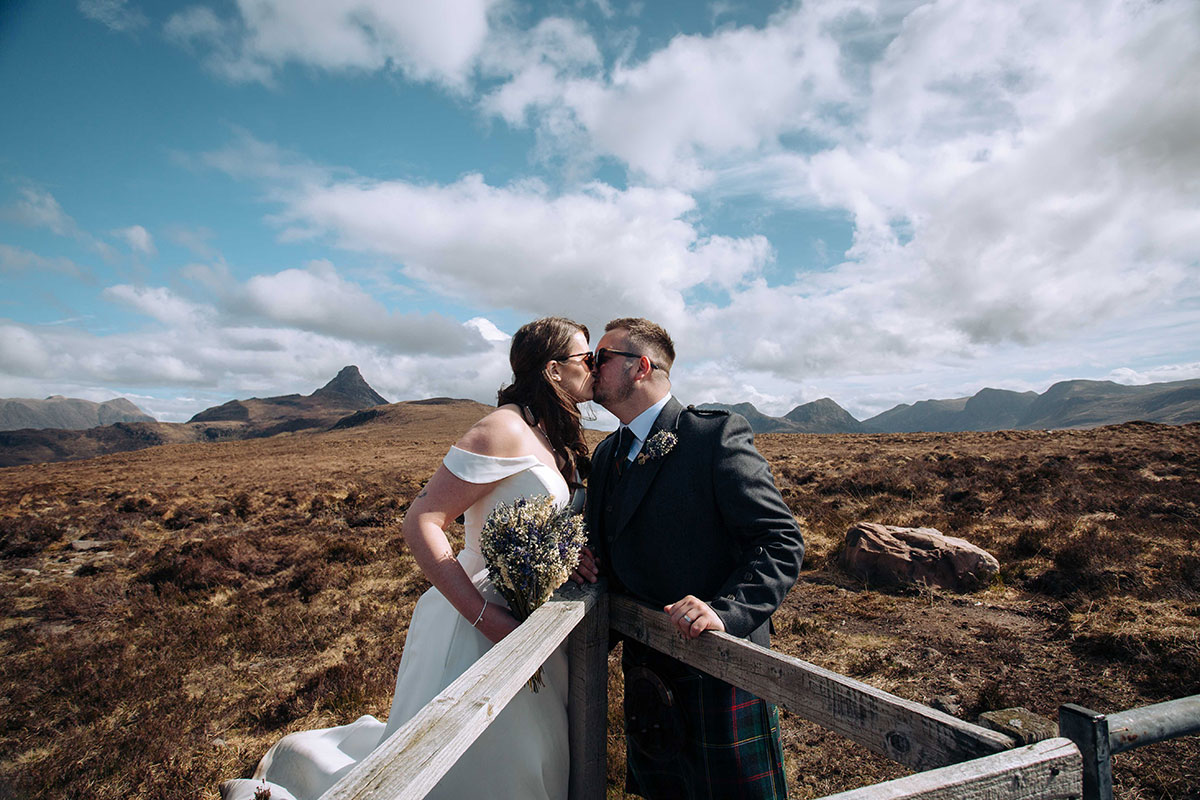 Bride and groom share a kiss beside a wooden fence with striking Highland peaks and open moorland in the background.