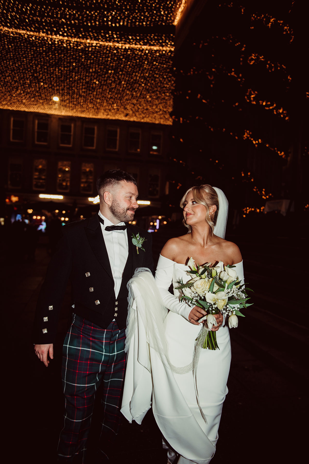 Bride and groom walking together at night on a city street, the bride holding a bouquet of white flowers