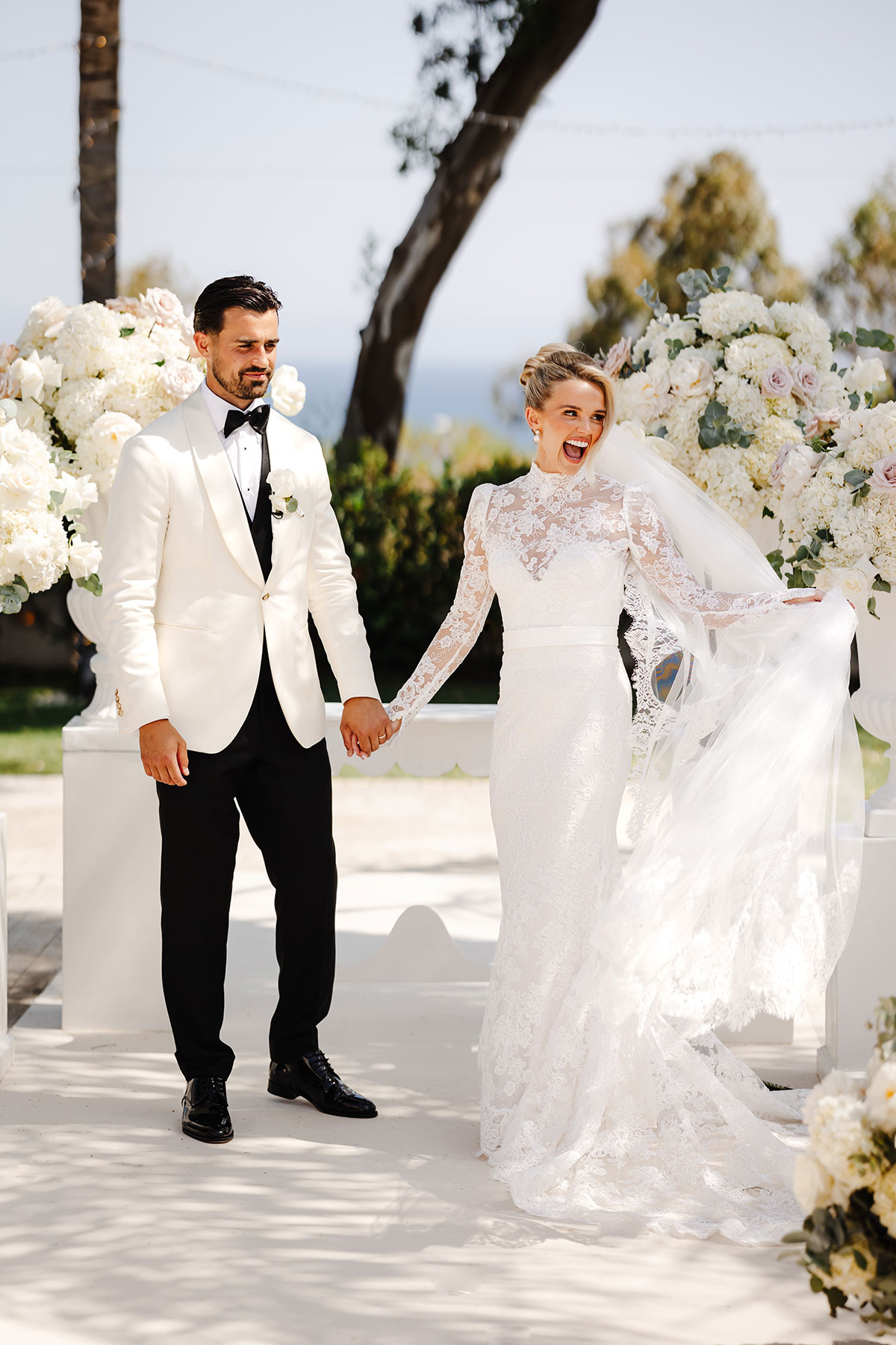 Bride and groom hold hands and smile during their outdoor wedding ceremony, surrounded by tall white floral arrangements and soft sunlight
