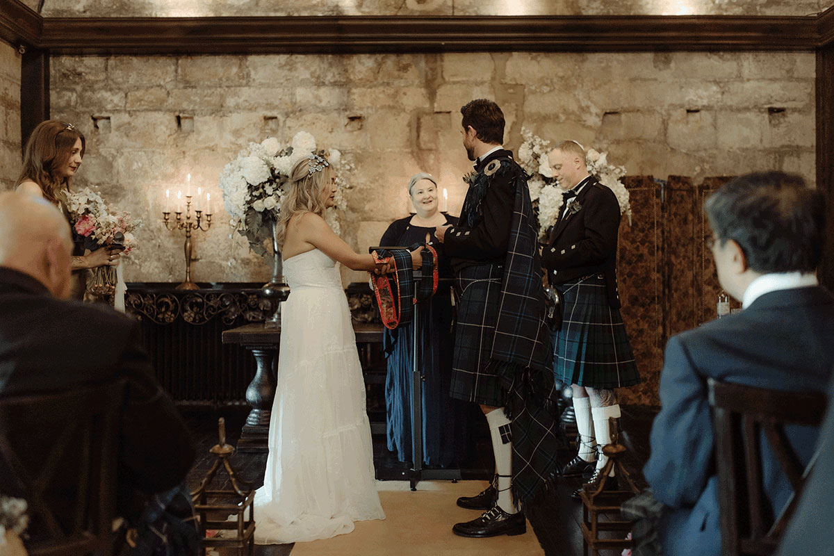 Scottish wedding ceremony inside Borthwick Castle featuring Fuze Ceremonies celebrant Yvonne leading a handfasting ritual surrounded by candlelight and stone walls