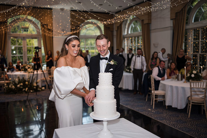 Bride and groom smile while cutting white three-tier cake