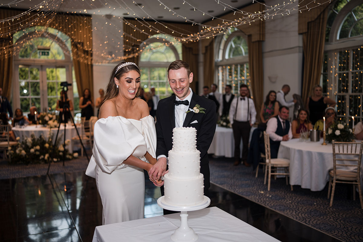 Bride and groom smile while cutting white three-tier cake