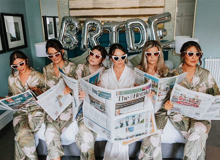Bride and bridesmaids with pyjamas and sunglasses on reading newspapers sitting on bed 