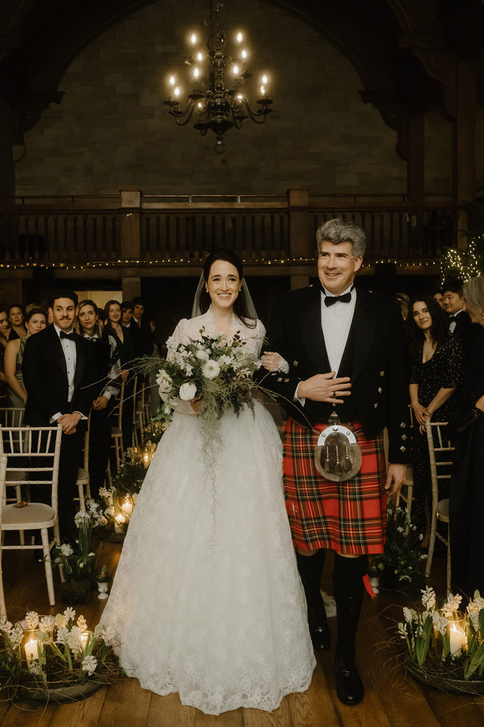 a bride walking down an aisle on the arm of a man wearing a red tartan kilt.