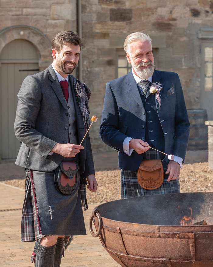 father and son wearing scottish wedding outfits toast marshmallows by the fire at rosebery steading