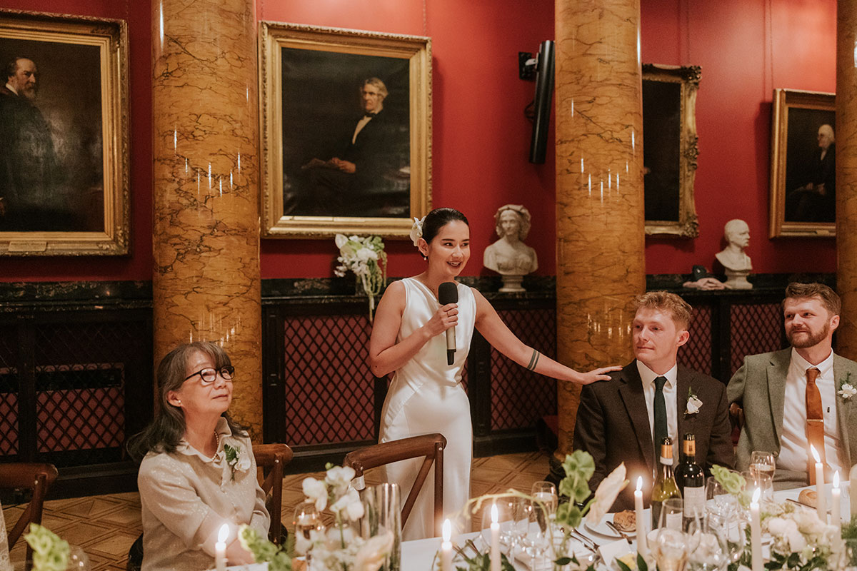 Bride giving wedding speech with microphone, standing beside groom at reception.