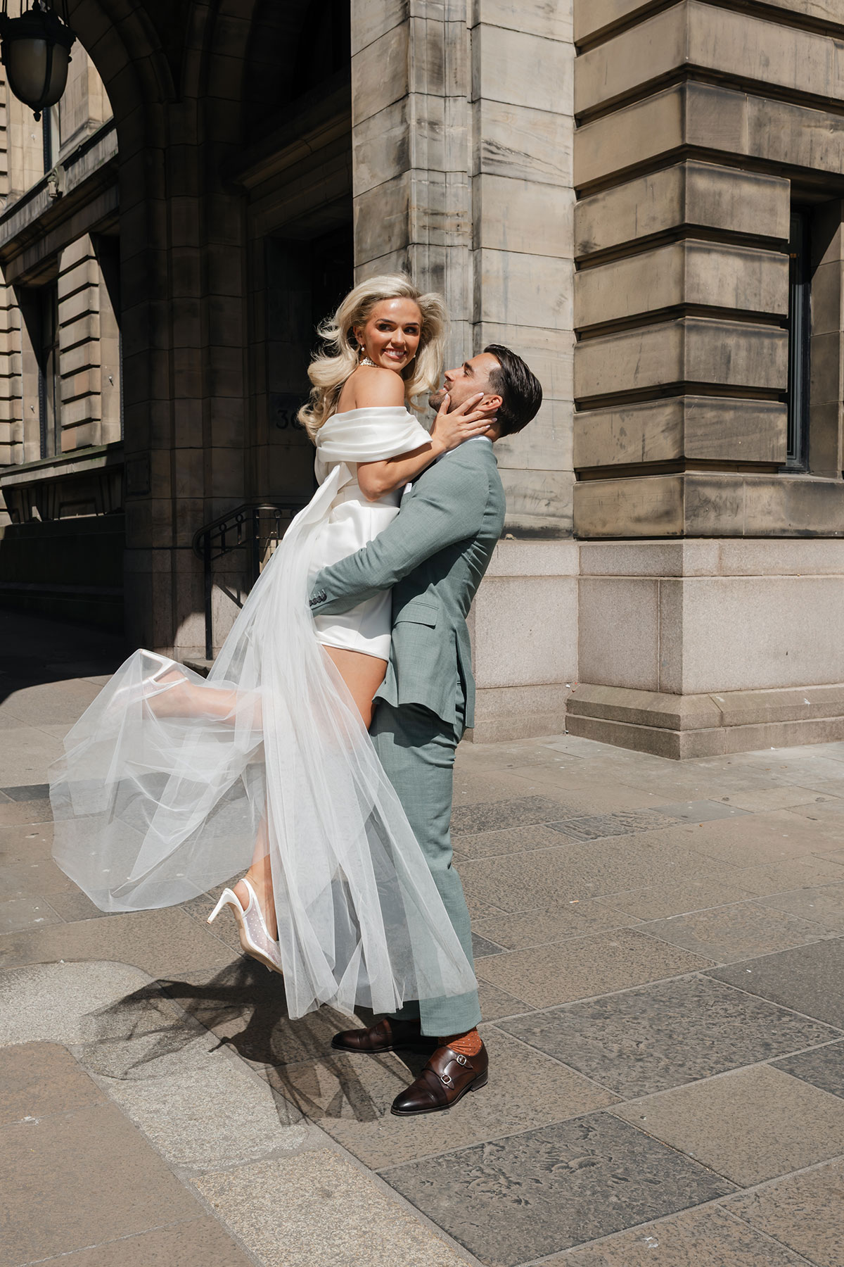 Groom lifts bride wearing short white dress with bow back and long tulle detail against sandstone city building backdrop