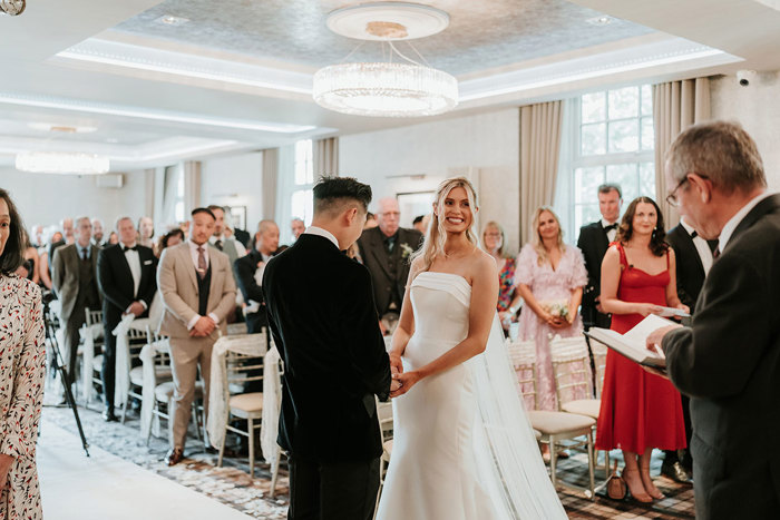 a bride and groom standing holding hands during their wedding ceremony at busby hotel as rows of standing guests watch on