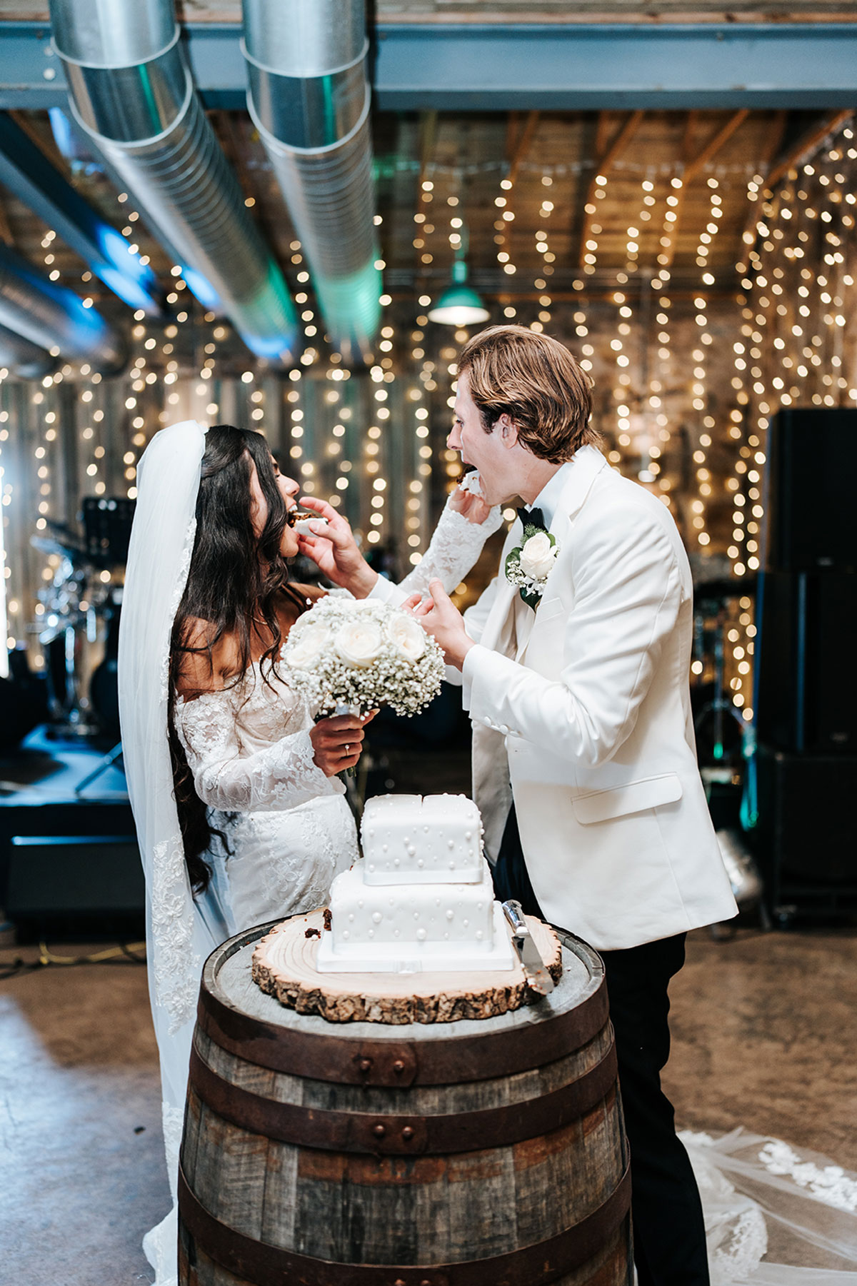 Bride and groom feeding each other cake beside a white tiered wedding cake under fairy lights