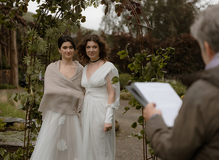 two brides standing during their wedding ceremony outside under a foliage-covered wire arch in a garden with old stone building in background. There is a celebrant in the foreground