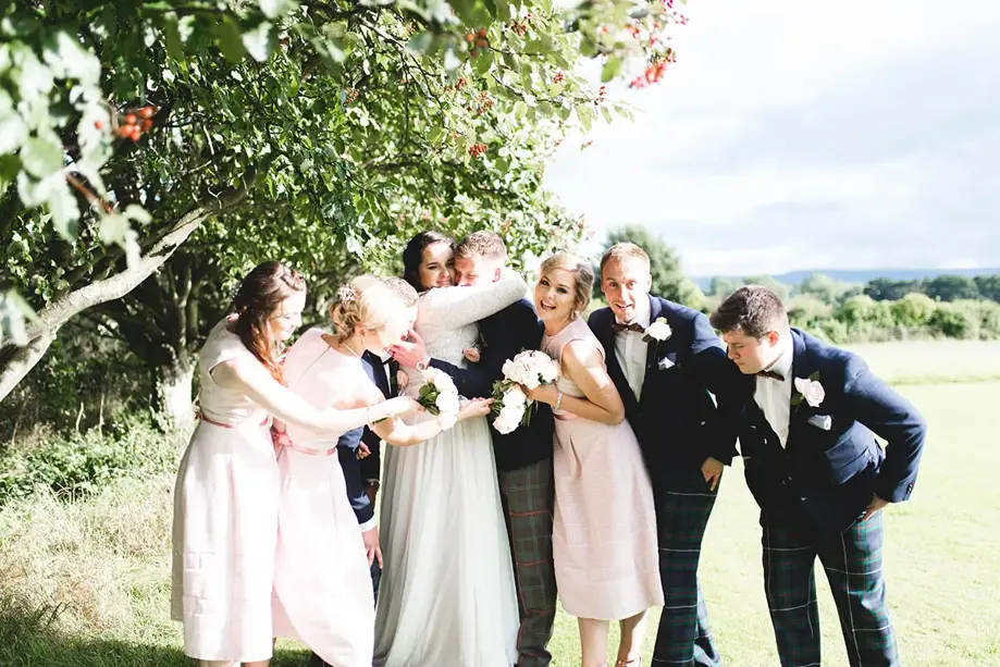 Bridal party and groomsmen laugh whilst bride and groom embrace in field