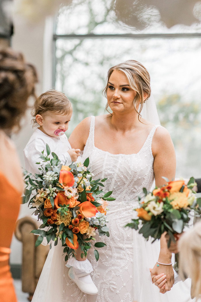 bride in beaded gown carries little girl dressed in white alongside an orange and green bouquet