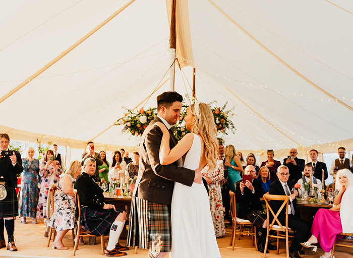 a bride and groom dance in a sailcloth marquee as gathered guests look on. A circular floral decoration is suspended from the ceiling behind them