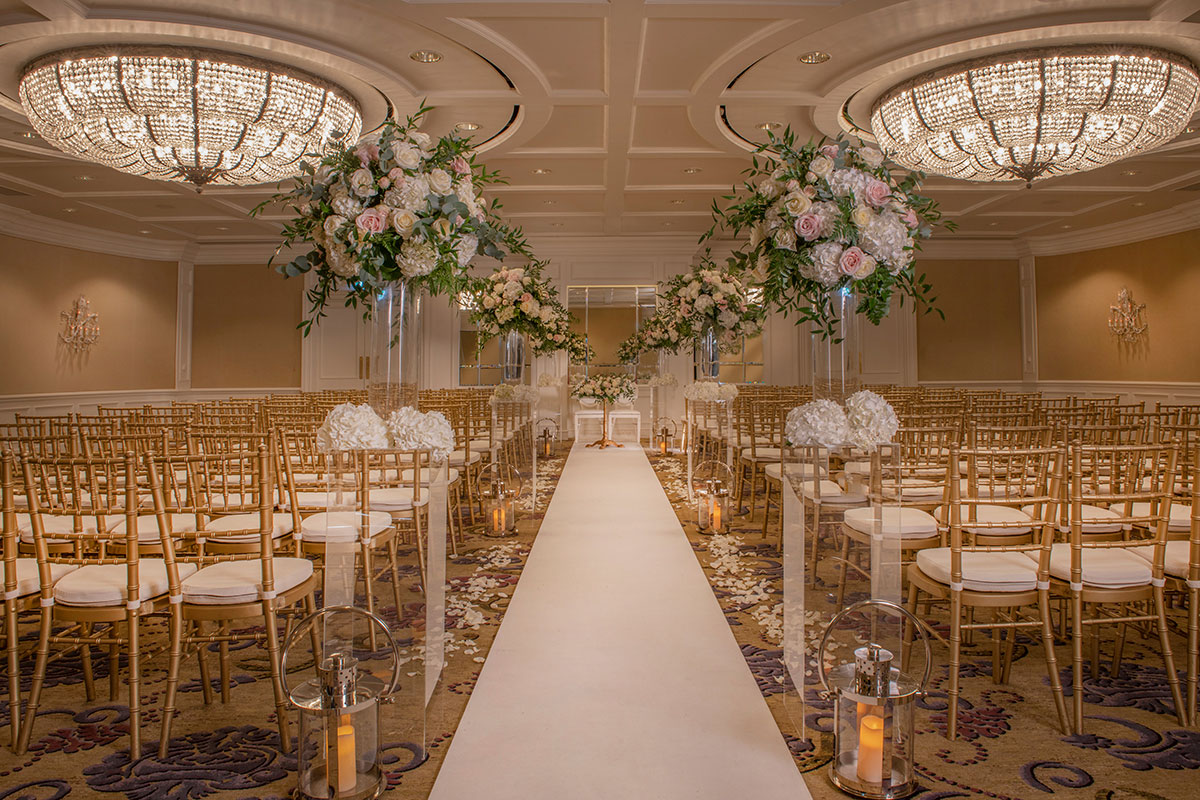 large golden patterned carpet room with white runner down the centre with rows of golden chairs with white cushions lined on either side with a few bouquets of white flowers and large pastel displays of flowers raised on clear stands