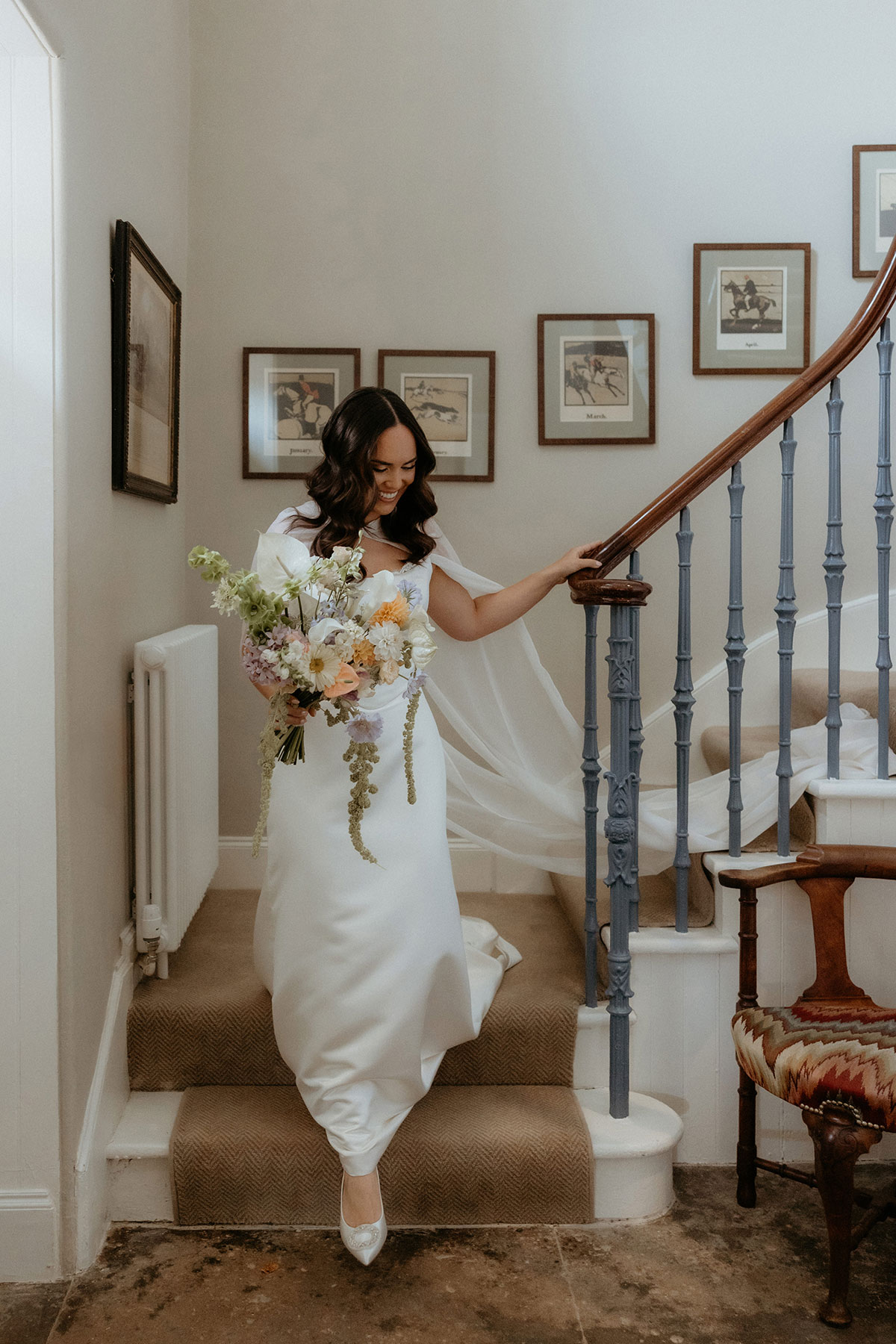 Bride descending staircase at Rosebery House, Midlothian, holding pastel bouquet and wearing modern satin wedding dress with cape.
