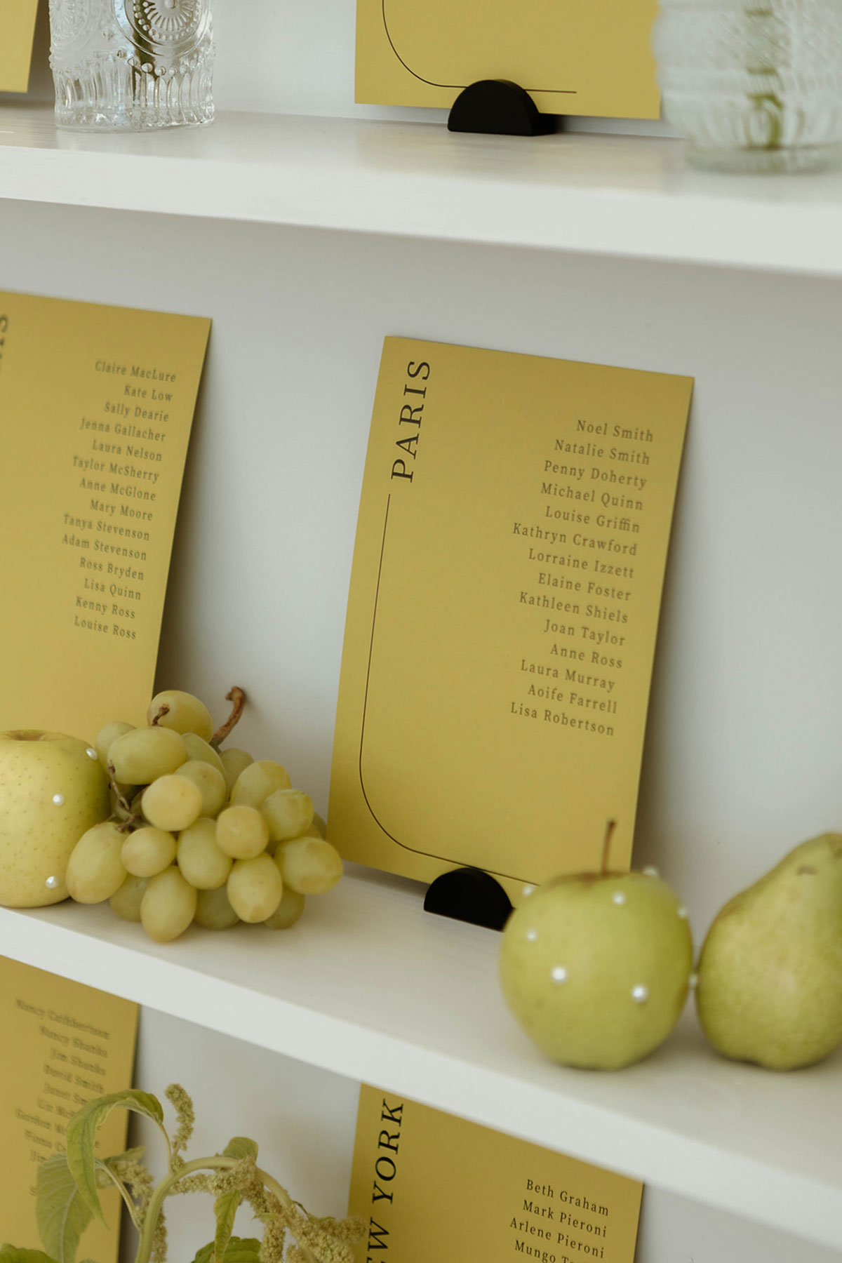 Yellow wedding seating plan cards on white shelving with green apples and grapes styling.