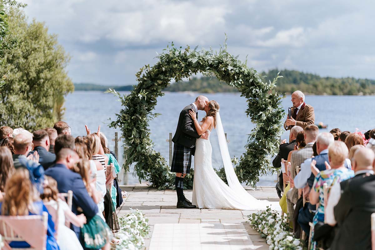 bride and groom kiss at top of aisle with circular arch and loch view behind them