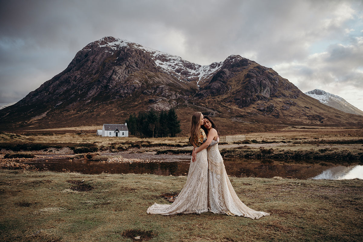 Couple embracing beside the river in Glencoe with a white cottage and mountain scenery behind them.