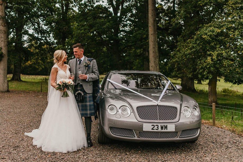A bride in a long dress and a groom in a kilt stand next to a silver Bentley with white ribbons attached to the bonnet