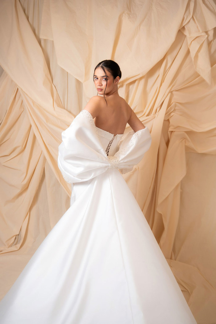 bride with dark brown hair wears strapless ballgown wedding dress and looks back over her shoulder to show off large bow feature on the back of her waist as she stands in front of a dark cream sheet backdrop
