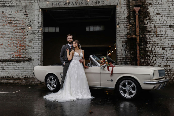 bride and groom stand in front of cream convertible car, bride reaching behind her and holding onto groom's label while bridal bouquet lies on the car's bonnet