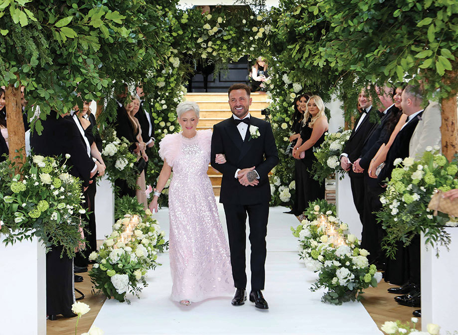 a groom wearing a smart black suit and bow tie walking down a white-carpeted aisle on the arm of a woman wearing a pink sparkly dress. There is tall greenery on either side of the aisle and green and white floral arrangements on the floor. Smiling guests stand either side looking on
