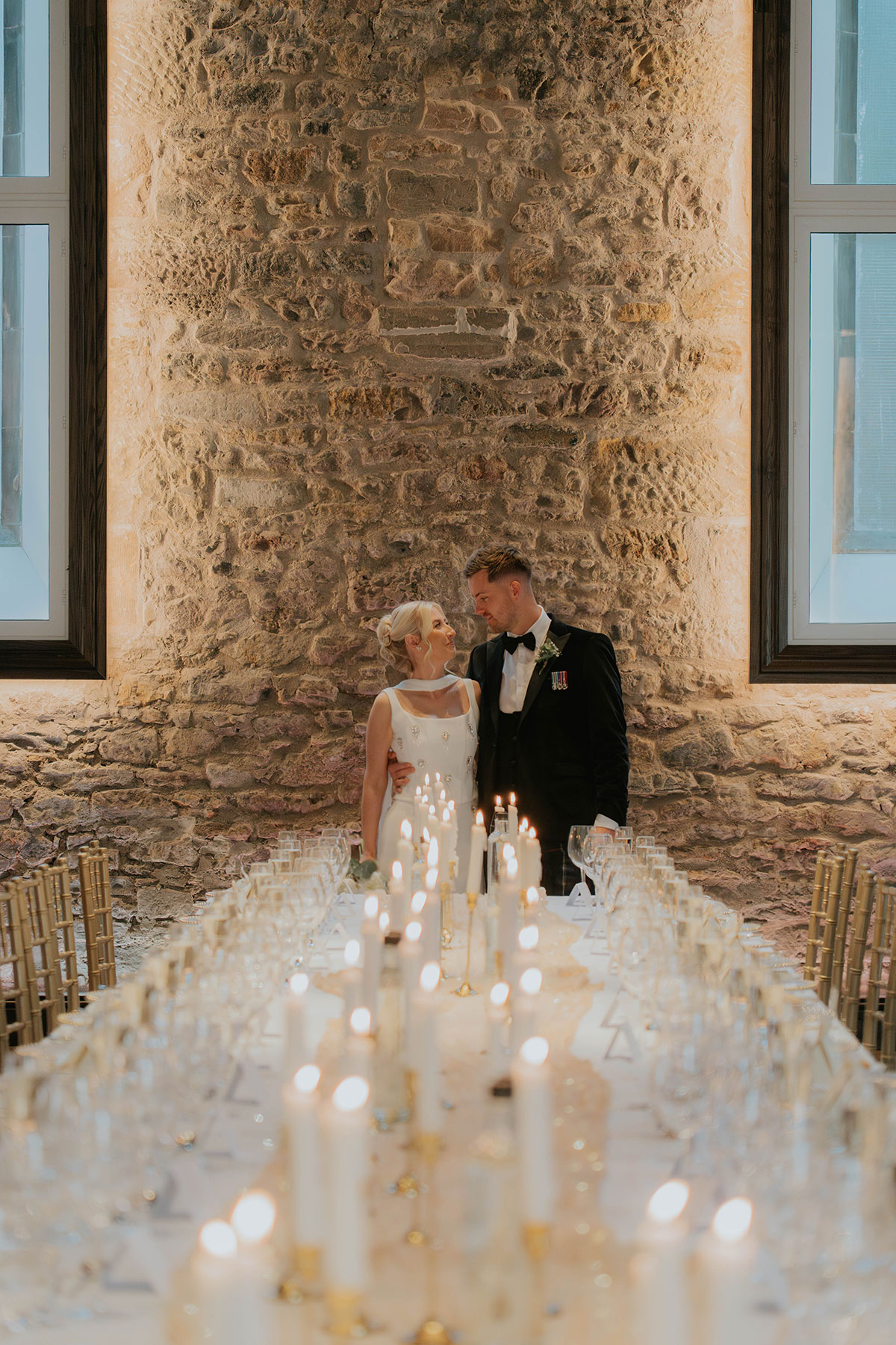 Bride and groom beside a candlelit wedding breakfast table in a historic stone-walled Edinburgh venue