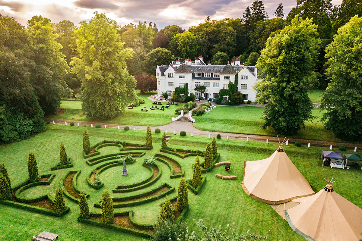 A large country house with manicured formal gardens and tents set on expansive grounds.