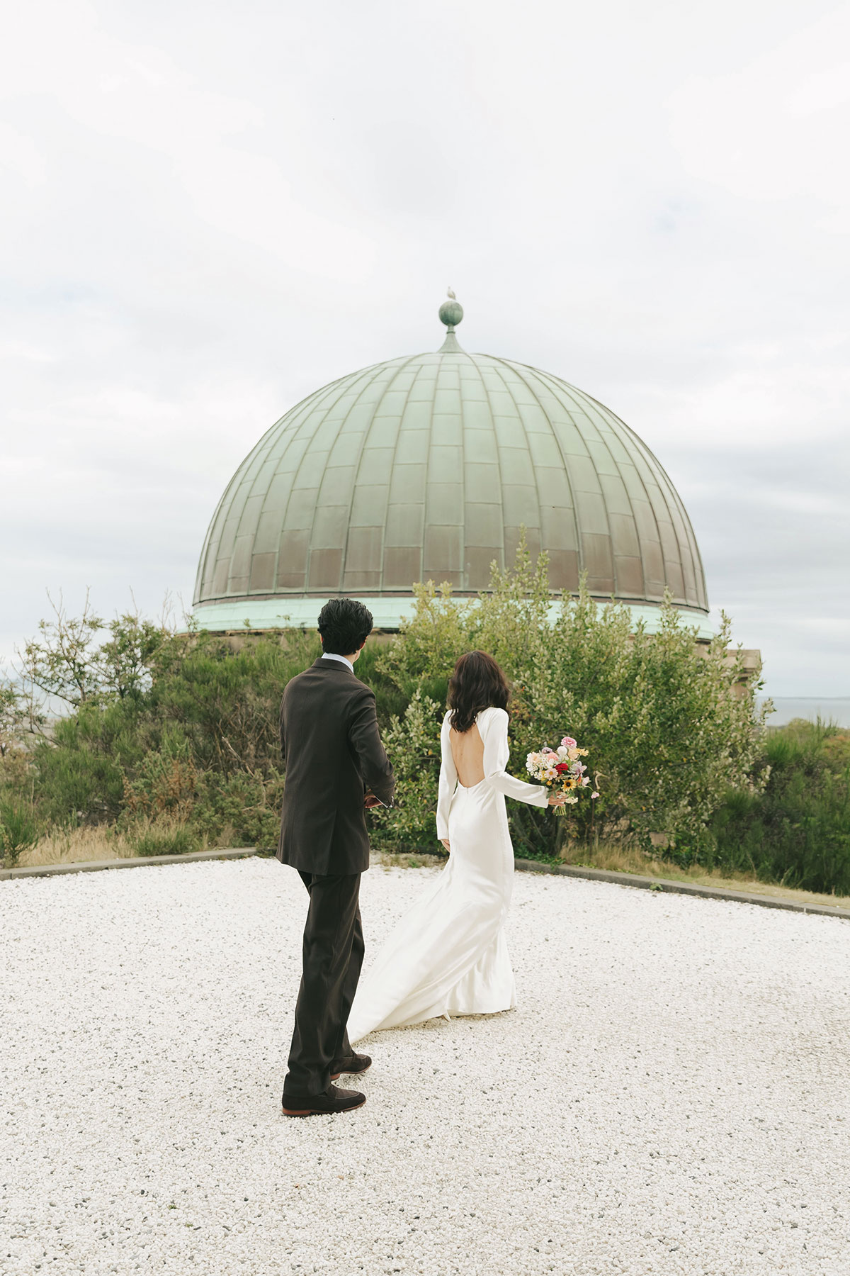 Bride and groom walking towards a domed observatory building, with the bride holding a bouquet and her backless gown flowing behind her