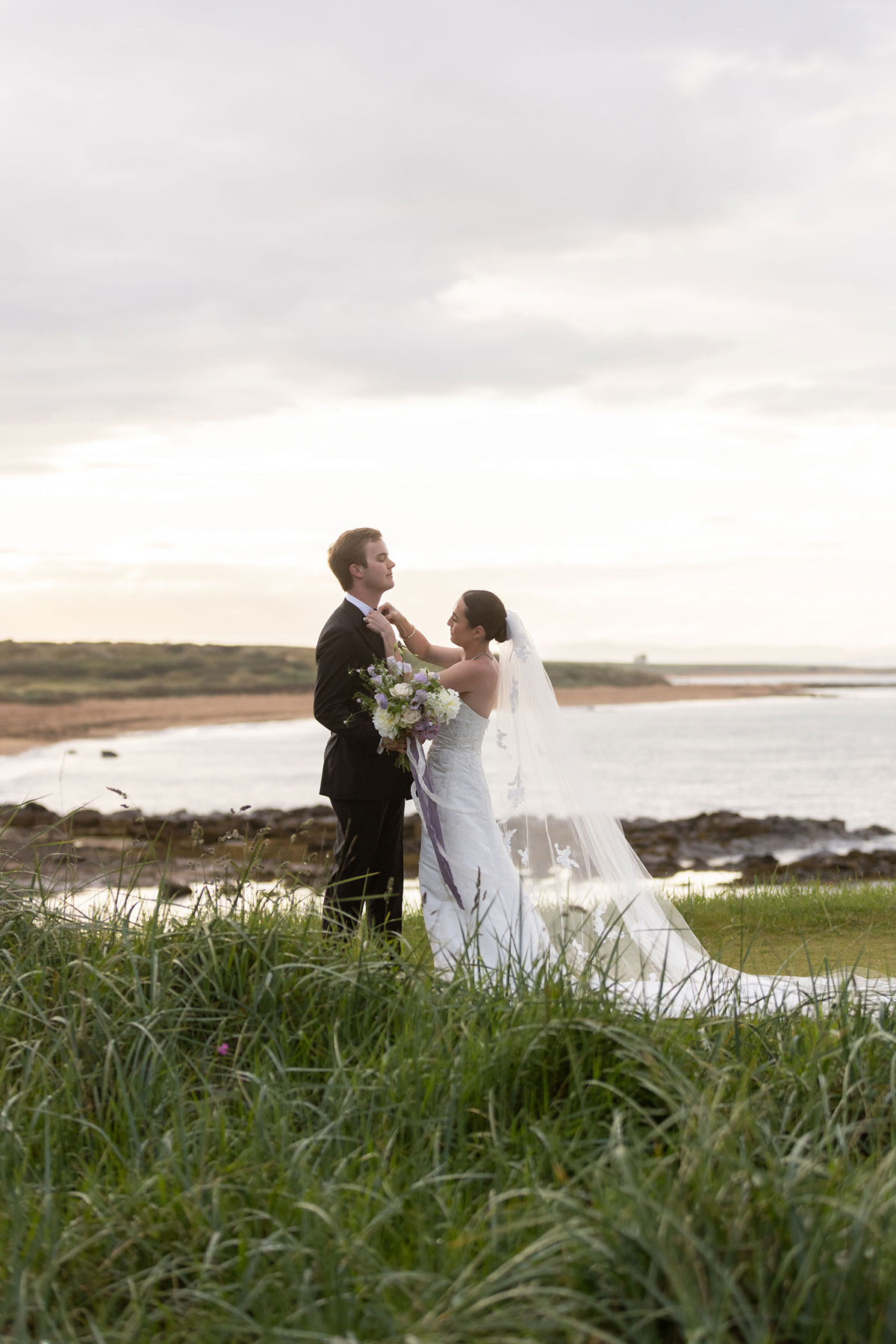 Bride and groom standing by the coast in Scotland, adjusting the groom’s jacket during their wedding portraits.