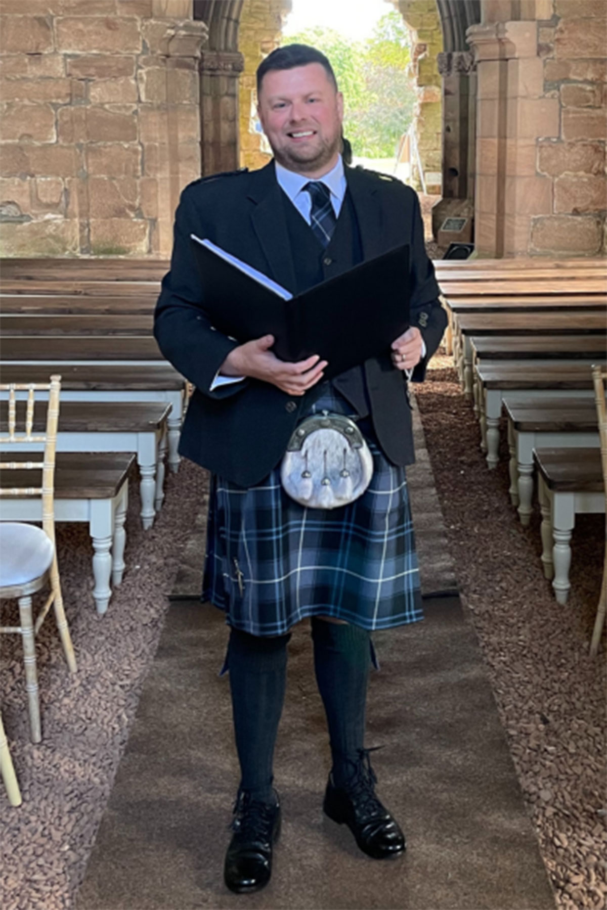 Scottish wedding celebrant standing in stone aisle wearing traditional kilt, holding ceremony folder at outdoor wedding venue