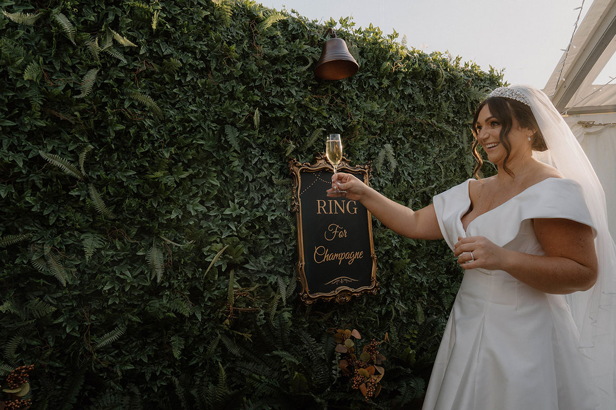 Bride ringing a “Ring for Champagne” bell against a foliage wall at a Cluny Castle wedding in Aberdeenshire.
