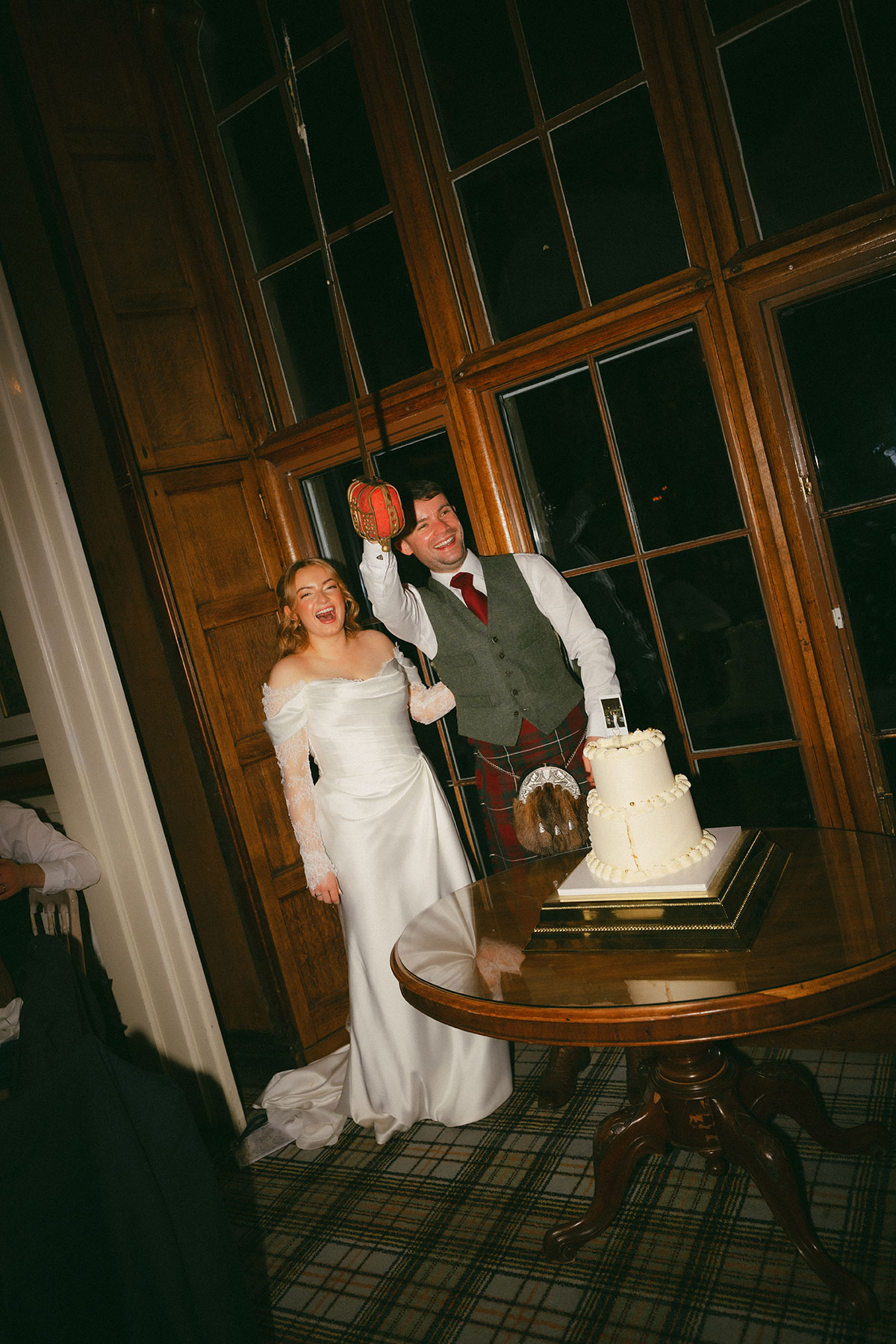 Bride and groom cutting wedding cake indoors at Scottish venue, groom in kilt and bride in lace-sleeve dress