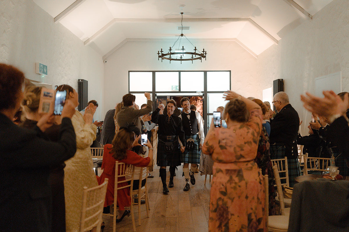 Two grooms make their reception entrance to applause from guests, both wearing Gunn tartan kilt outfits at Newhall Estate