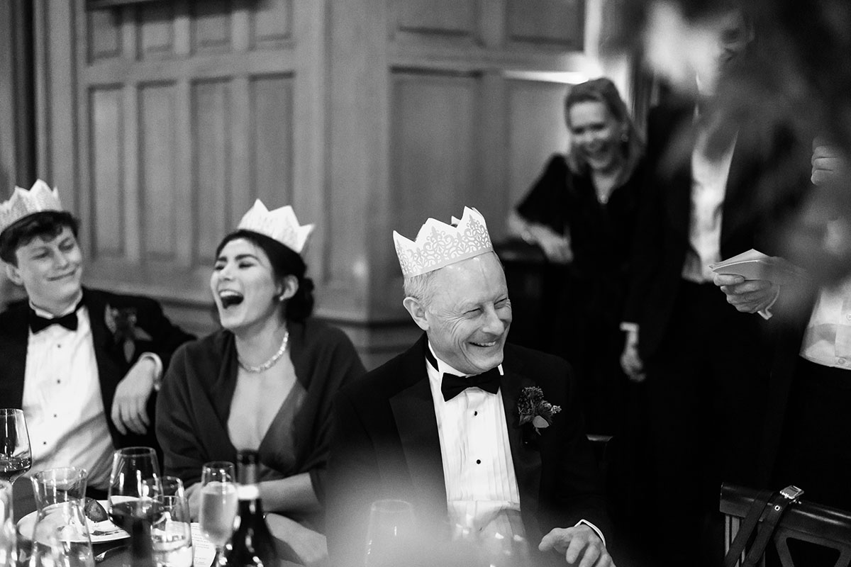 Wedding guests in paper crowns laughing during speeches at the candlelit reception.