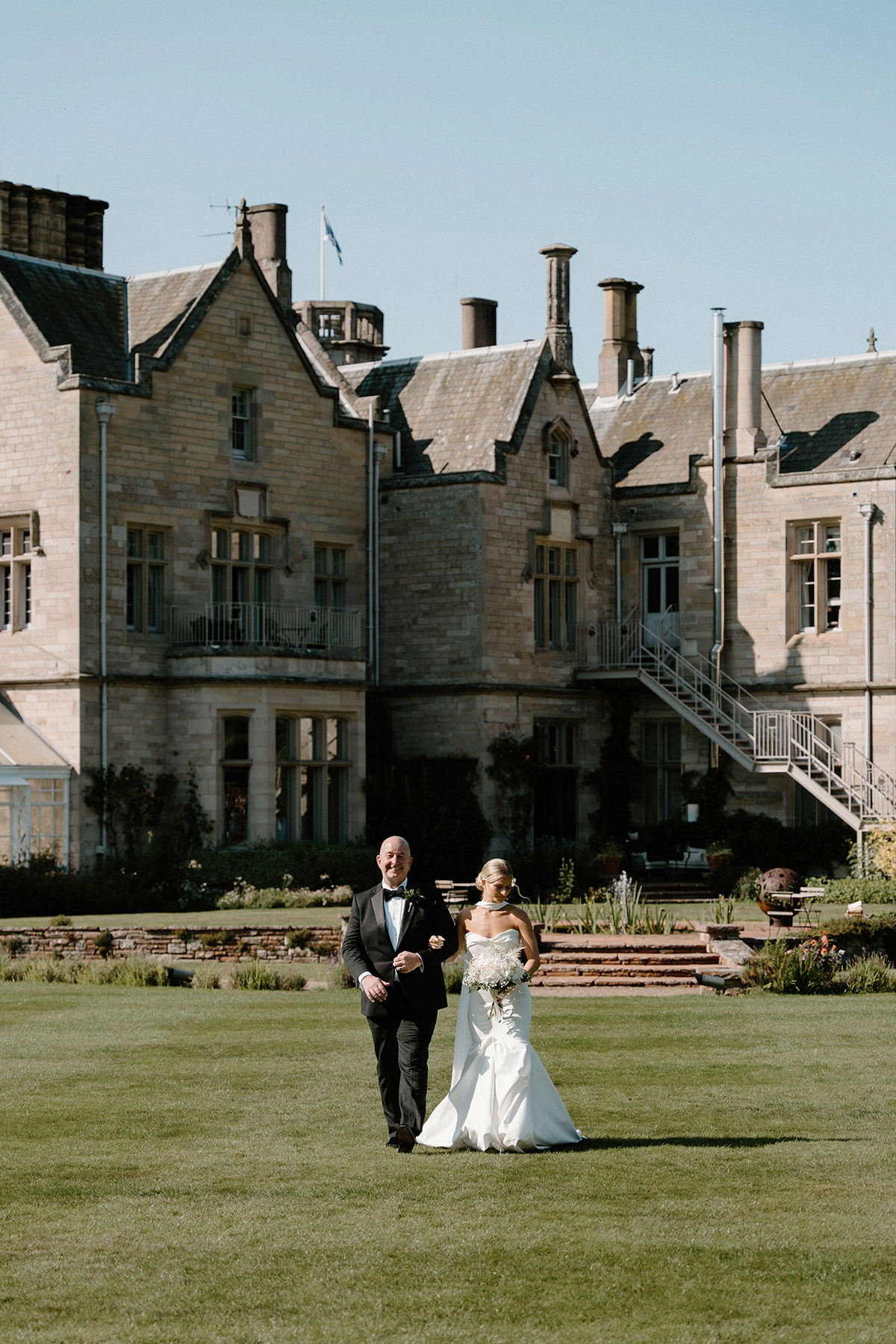 Bride walking across the lawn with her father outside a historic Scottish country house wedding venue before the outdoor ceremony