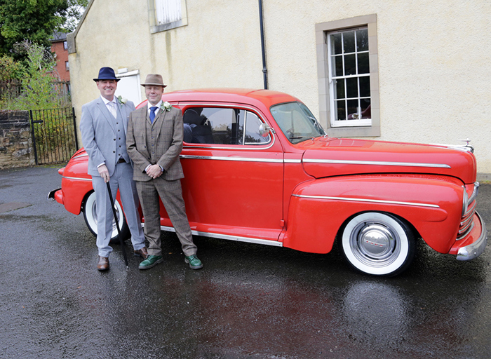 Two men in grey suits stand in front of a red car