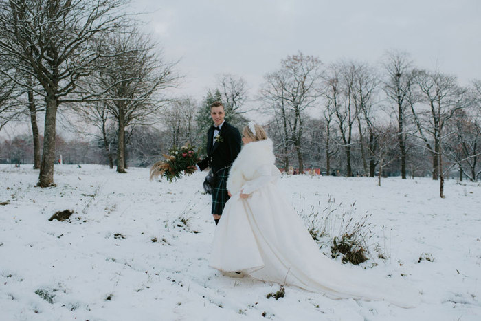 A bride wearing a white dress and white fur wrap holding hands with a groom wearing a dark kilt walking in a park in the snow