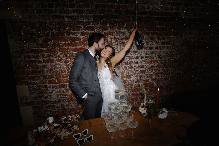 with his arm around her waist, groom kisses bride on the cheek as she smiles widely and lifts a bottle of champagne up in the air, a champagne tower sits on the table in front of them